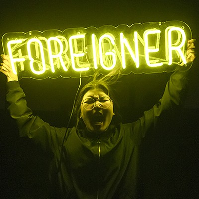 A woman screams in a darkened room, holding up a yellow neon sign that reads "FOREIGNER"