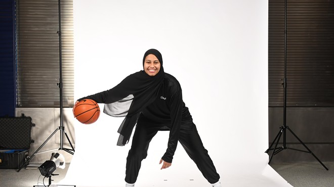 A tall woman with brown skin smiles while dribbling a basketball in loose-fitting activewear and an athletic-cut hijab