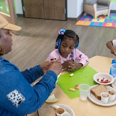 Two young kids with braided hair and dark skin listen as a woman peels a banana at a table with sandwiches on plates