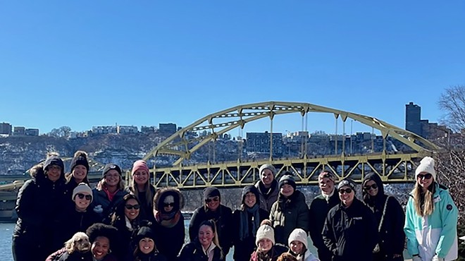 A group of women in coats and hats and three medium-sized dogs stand in the sun with snowy Mt. Washington and a large arch bridge in background
