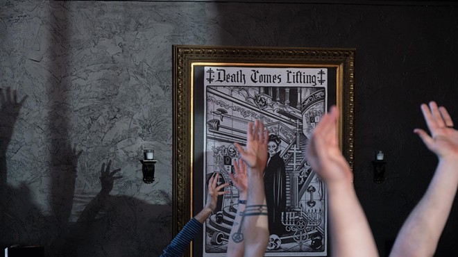 Tattooed arms are extended near a Death Comes Lifting print in a gilded frame