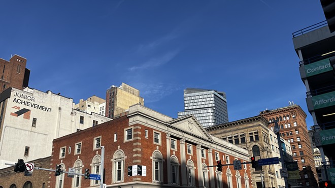 A low brick building with Neoclassical details is illuminated by the sun with skyscrapers rising behind it.