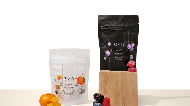 Photo of two bags of evn THC gummies, white bag on the left is Sativa variety with citrus imagery on it, black bag on the right is Indica variety with berry imagery. Bags are staged in front of a white background on a beige foreground, with fruits and gummies scattered around them.