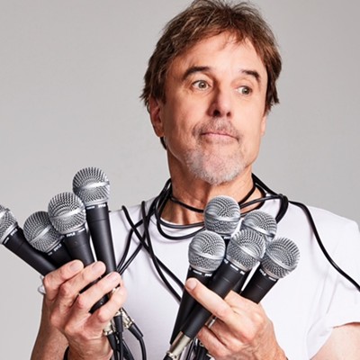 A man in a white T-shirt with a goatee holds fistfuls of microphones while grimacing