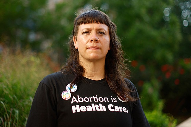 A woman poses in front of a field for a portrait wearing a black shirt that has written on it, "Abortion is Health Care." and a couple pins on her shirt, one of which is a logo for Planned Parenthood.