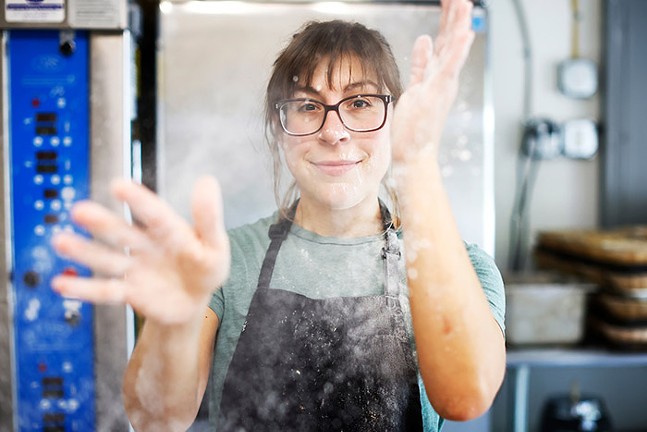 Woman with brown hair and dark-rimmed glasses is wearing an apron as she reaches out her hands in front of her and spreads out white flour crumbs to the camera