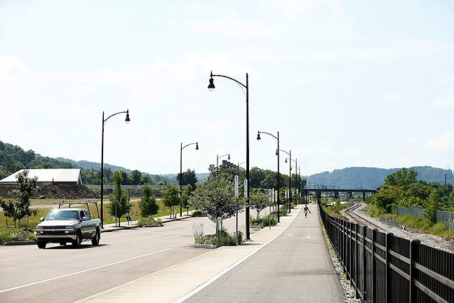 A green space sits beside a highway, walking and bike trail, and train tracks