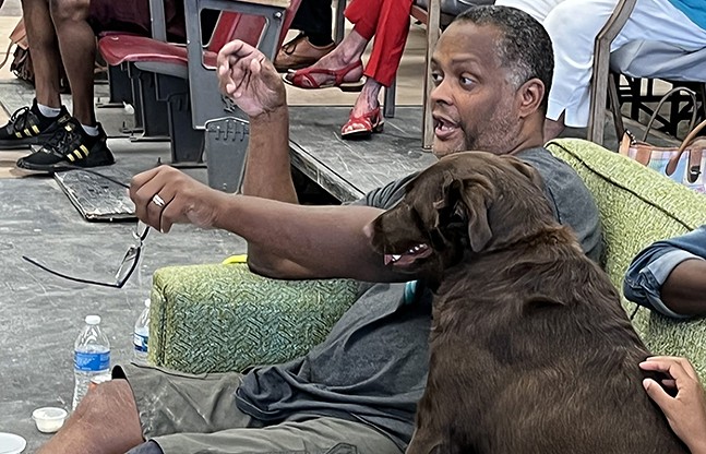 A man sits on a chair with dog pointing his hound out and speaking