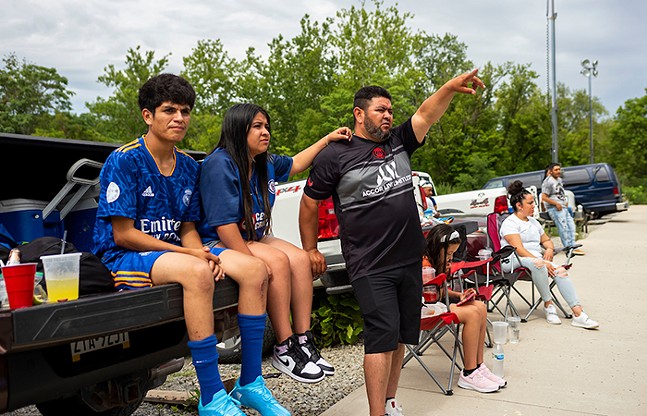 Two young people sit on the back of a truck watching a soccer game among others.