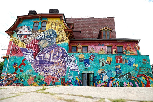 A colorful mural on the side of the building features a large painting of a man in a beret, surrounded by a collage containing people, illustrations, and the front covers of playbooks