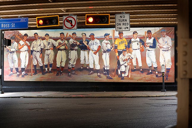 A long horizontal mural of 14 baseball players is painted on the side of a building behind street signs