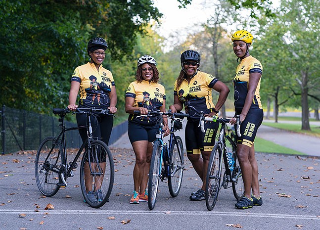Four smiling Black women wearing bicycle helmets stand next to bikes