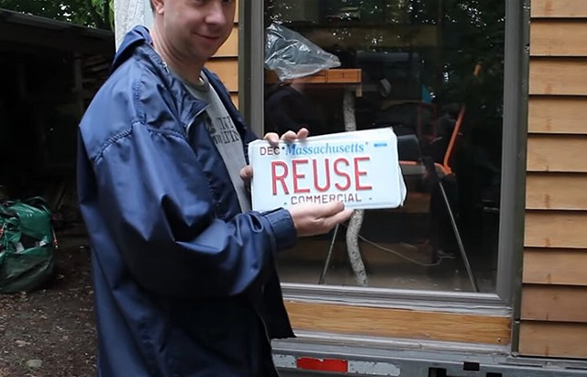 A smiling man in a rain jacket stands in front of a tiny house and holes up a Massachusetts license plate that says "REUSE"