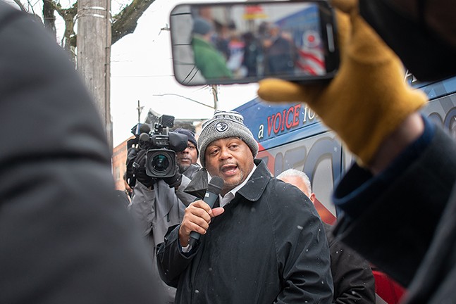 A person holds up a cell phone in the foreground, and a man holds up a video camera in the background, both recording a man who's wearing a Pittsburgh Steelers hat and talking into a microphone