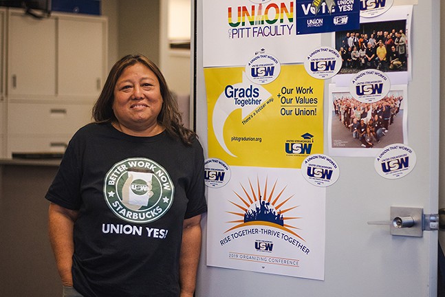 A smiling woman wears a "Union Yes!" T-shirt as she leans against a door covered in pro-union signs