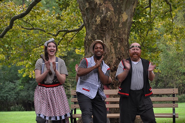 Three people in old-timey clothes laugh and clap their hands in front of a park bench and large tree