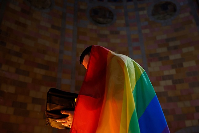 A person reading the bible with an LGBTQ flag draped over their body, hiding their identity