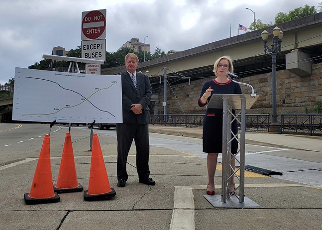 A man stands beside a map held up with three orange traffic cones. To his left, a woman stands talking at a podium. Both are pictured at a city bus stop.