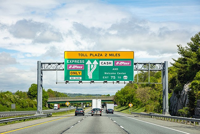 Cars and trucks drive away from the camera on a highway. Trees are on both sides of the road, and a sign goes over the highway saying, "Toll Plaza 2 Miles"