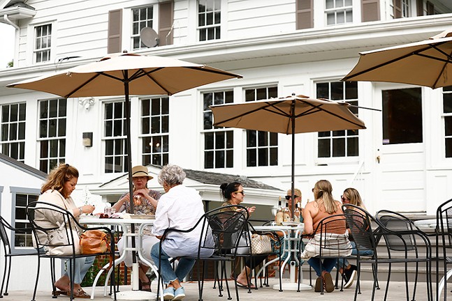 Two outdoor table fulls of women drinking under patio umbrellas in front of a large white building with lots of windows