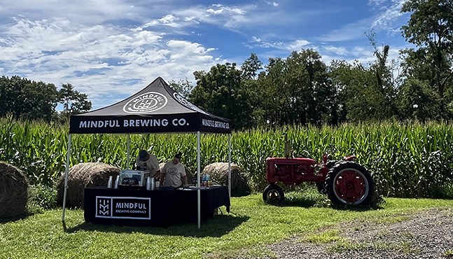 A picturesque view of a cornfield, hay bales, and a tractor under a blue sky. On the grass in the foreground, two people serve beer underneath a tent that reads "Mindful Brewing Co."