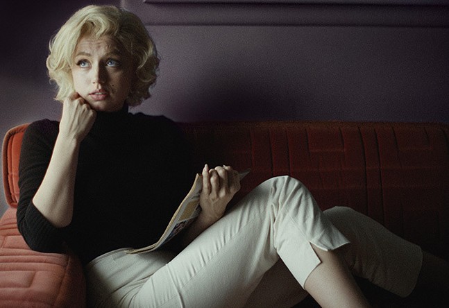 Actress Ana de Armas, wearing a black turtleneck sweater and tan capris, poses on a couch in her role as the late Marilyn Monroe in the movie Blonde.