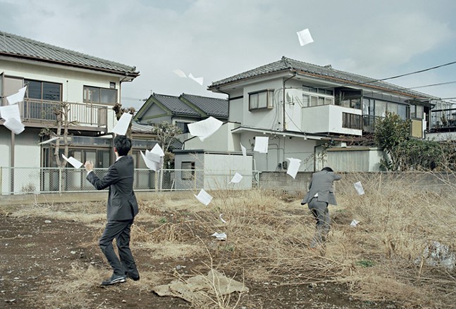 Two people in business suits chase after papers blowing in the wind.