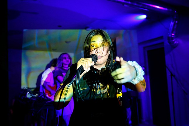 A woman holds a mic and points at the camera