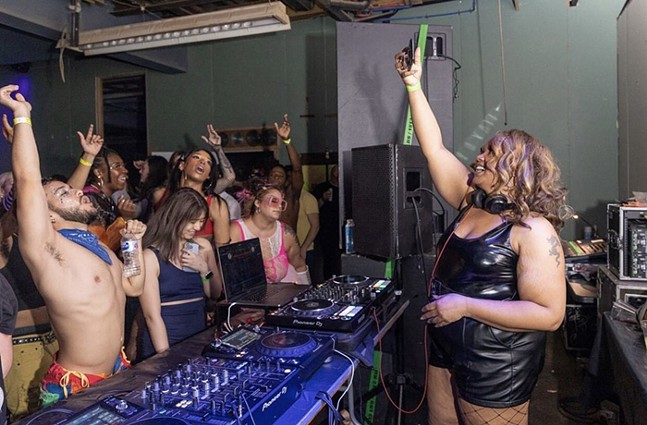 A Black female DJ holds up her smartphone to photograph an energetic dance crowd.