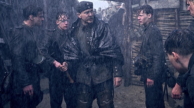 A group of World War One soldiers stands around in a heavy storm, their hair and uniforms slick with rain.