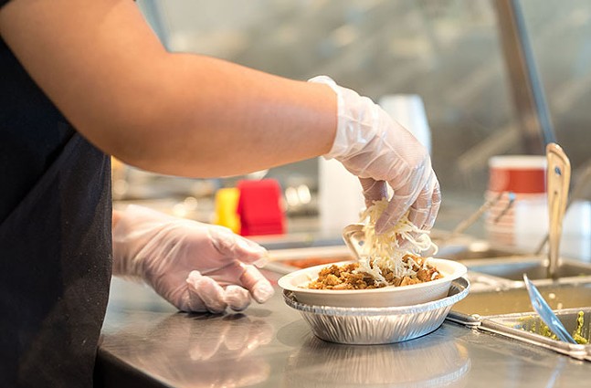 A close-up shot of a restaurant worker wearing latex gloves as they sprinkle cheese on a burrito bowl.