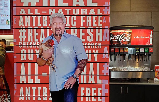 A large photo of Guy Fieri holding a chicken sits in a fast-food restaurant dining room.