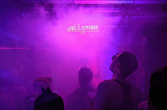 Fog blurs a neon sign that says Jellyfish with silhouettes of people dancing in the foreground.