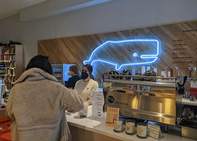 A woman wearing a black face mask serves a customer at a coffee counter as a blue neon outline of a whale glows in the background.