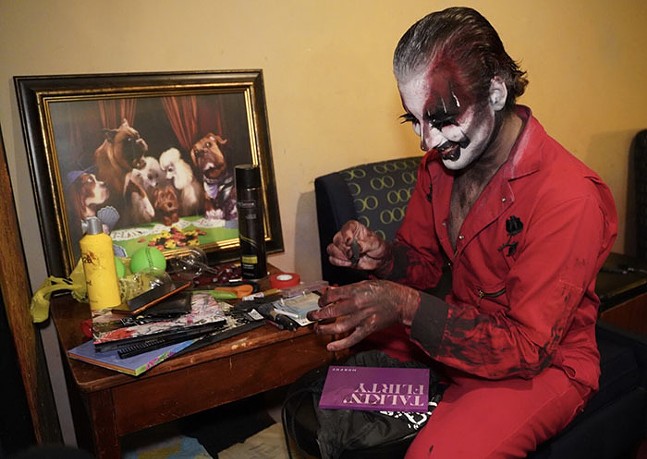 A drag performer with slicked-back hair, wearing a red outfit and face paint, smiles as they do their makeup.
