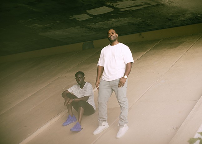 Two Black men, both wearing white T-shirts, pose on the slope under and overpass.