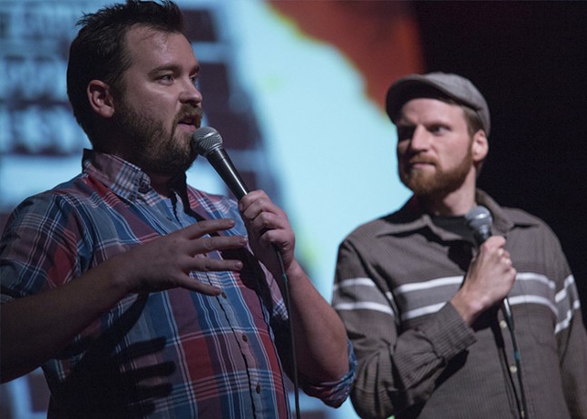 Two white men, one wearing a plaid shirt, another with a reddish beard and wearing a flat cap, hold microphones as they perform on stage