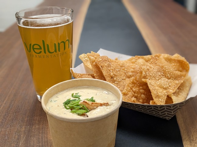 A tall beer in a glass labeled "Velum" sits in the background of a takeout order of tortilla chips and blanco cheese dip.