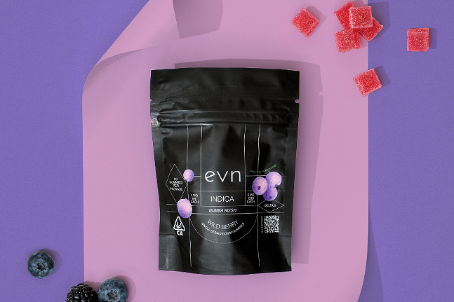 Photo: black bag of evn indica gummies with wild berry flavor, background is different shades of purple with red gummies and purple mixed berries scattered in frame
