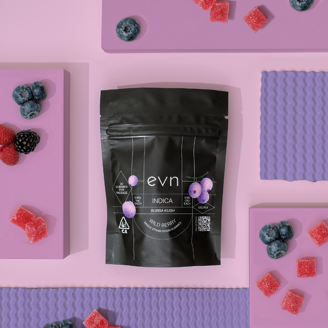 Photo of black bag of evn Indica gummies in the flavor Wild Berry. Background is abstract shades of purple with gummies and berries scattered around the bag.