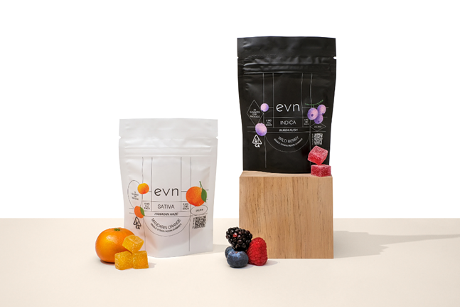 Photo of two bags of evn THC gummies, white bag on the left is Sativa variety with citrus imagery on it, black bag on the right is Indica variety with berry imagery. Bags are staged in front of a white background on a beige foreground, with fruits and gummies scattered around them.