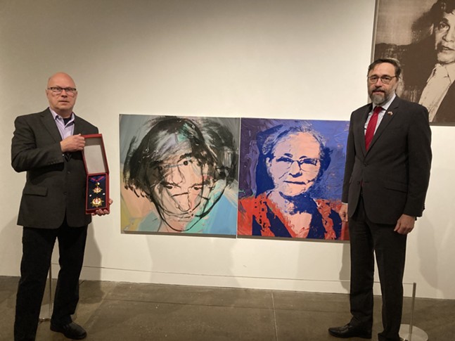 Two middle-aged men in tailored suits, one of whom holds a large medallion in a frame, stand near Andy Warhol's portraits of Marilyn Monroe and his mother.