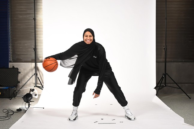 A tall woman with brown skin smiles while dribbling a basketball in loose-fitting activewear and an athletic-cut hijab