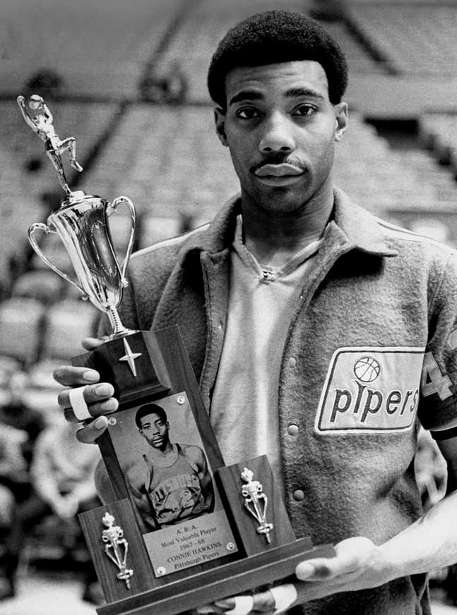 A man in a Pittsburgh Pipers jacket with a short afro holds an ornate trophy with his photo on it