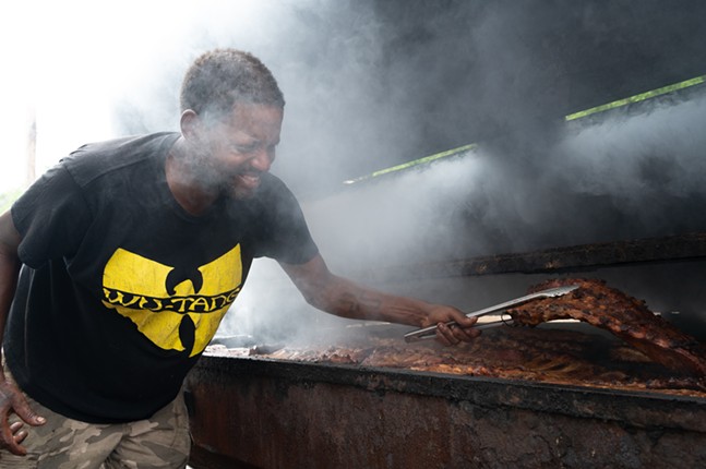 A man with short hair in a Wu-Tang T-shirt lifts a charred rack of ribs from the grill of a smoker