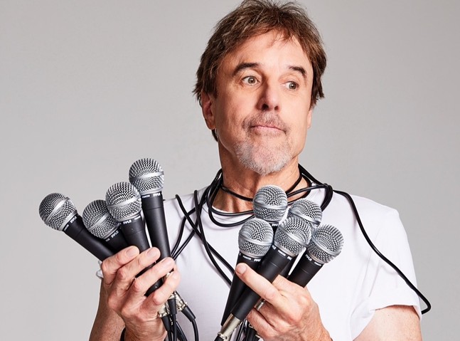 A man in a white T-shirt with a goatee holds fistfuls of microphones while grimacing