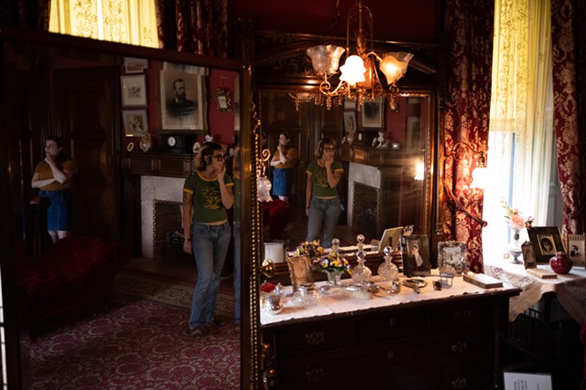 Two young women in casual clothes with tattoos stand near a dresser covered in cut-glass perfume bottles