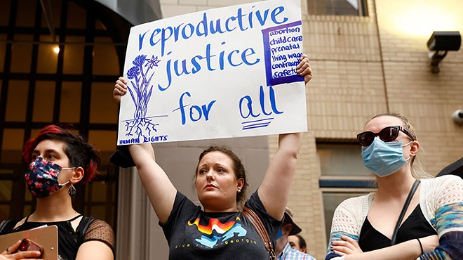 A woman holds up a sign that reads, 
"reproductive justice for all" in between two other people wearing masks with their arms crossed
