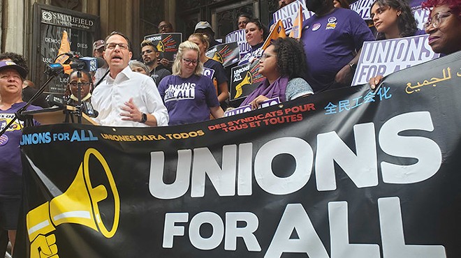 A man in a white shirt and glasses, backed by a large crowd of people, talks into a microphone in front of a huge black vinyl sign with an illustration of a yellow megaphone beside text that says "Unions for All" in multiple languages.