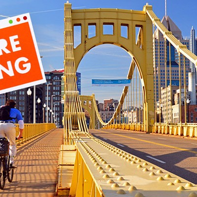 Person riding a bike across a yellow bridge cycling towards Pittsburgh city skyline. A sign that says "We're hiring" is Photoshopped on top of the photograph.